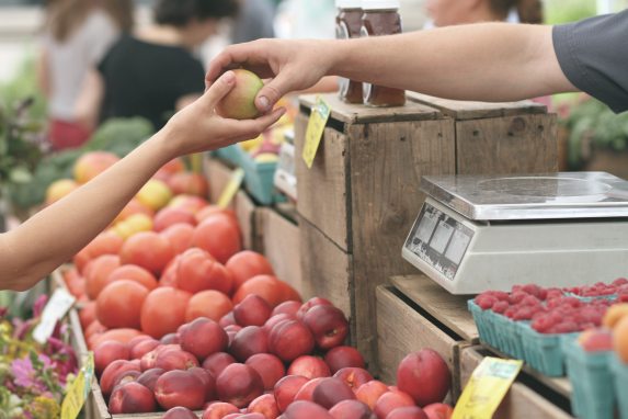 close up of two peoples' arms at a fruit stand; one person is handing an apple to the other