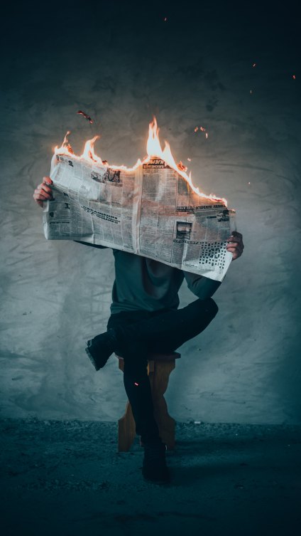 a person is sitting cross-legged while reading a newspaper that is on fire; the person's face is hidden