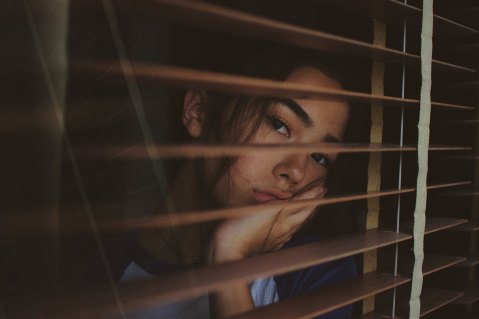 young girl in a dark room rests her face in her hand as she looks out between the slats of window blinds