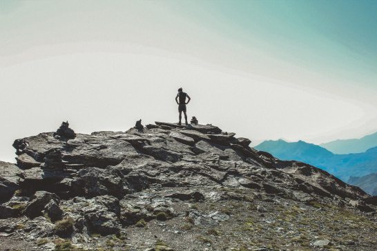 One person stands in silhouette on top of a rocky cliff with clear sky in the background