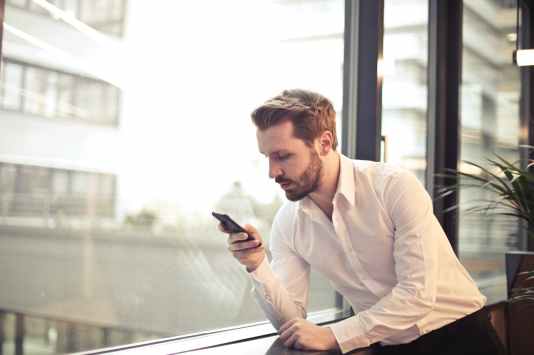 man with a beard is leaning against a glass window and reading from a mobile device